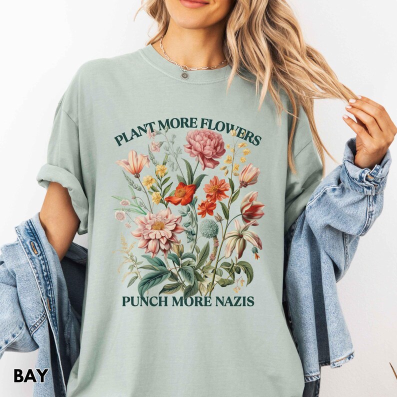 Op de afbeelding: Lichtgroen T-shirt met een bloemenontwerp met roze, rode en gele bloemen. De tekst "PLANT MORE FLOWERS" staat boven de bloemen en "PUNCH MORE NAZIS" eronder. Het shirt is gemaakt van een zacht materiaal.