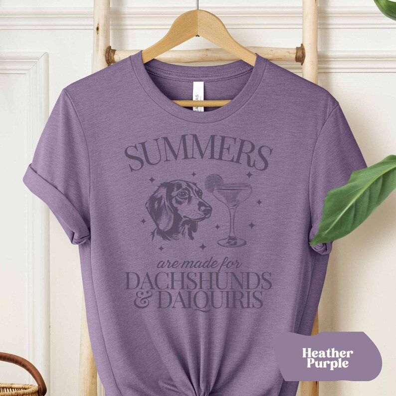 May include: A purple t-shirt with the text "SUMMERS are made for  DACHSHUNDS & DAIQUIRIS" and a graphic of a dachshund and a cocktail glass.