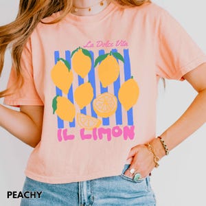 May include: Peach-colored t-shirt with a lemon graphic. The design includes yellow lemons, blue stripes, and the text "La Dolce Vita" and "IL LIMON". The word "PEACHY" is printed at the bottom left.