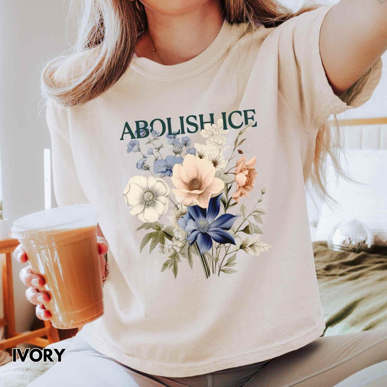 May include: Ivory-colored t-shirt with the text "ABOLISH ICE" in green above a floral design. The floral arrangement features white, peach, and blue flowers with green leaves. The word "IVORY" is printed at the bottom.