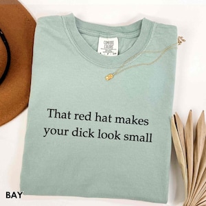 Może przedstawiać: Jasnozielona koszulka Comfort Colors z czarnym napisem "That red hat makes your dick look small". Złoty naszyjnik jest zarzucony na koszulkę. Słowo "BAY" jest wydrukowane w lewym dolnym rogu.
