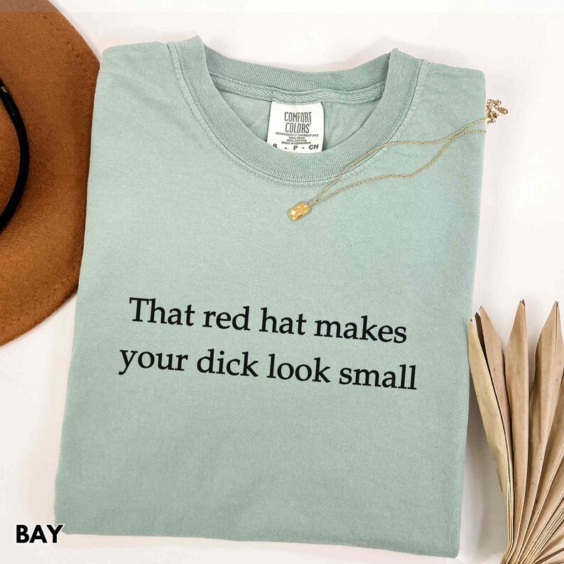 Może przedstawiać: Jasnozielona koszulka Comfort Colors z czarnym napisem "That red hat makes your dick look small". Złoty naszyjnik jest zarzucony na koszulkę. Słowo "BAY" jest wydrukowane w lewym dolnym rogu.