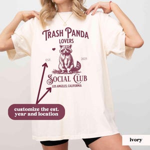 May include: A white t-shirt with a burgundy graphic of a raccoon with the text "Trash Panda Lovers" and "Social Club" with the text "Los Angeles, California" below. The text "Est." and "2025" are above the raccoon graphic.