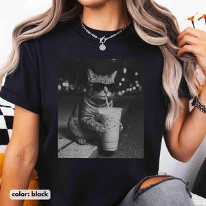 May include: Black t-shirt featuring a grayscale image of a cat wearing sunglasses and drinking from a cup with a straw. The cat is sitting on a curb with a blurred background of city lights. The text "color: black" is at the bottom.