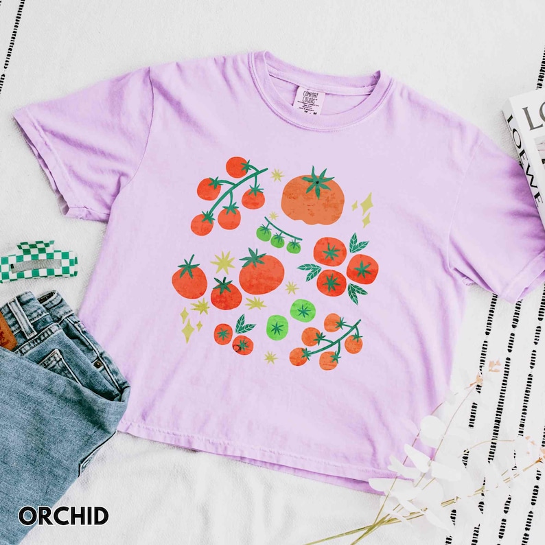 Puede incluir: Una camiseta de color lila claro con un dise&ntilde;o de tomate. El dise&ntilde;o presenta tomates naranjas y verdes con hojas verdes y estrellas amarillas. La palabra "ORCHID" est&aacute; impresa en la parte inferior de la imagen.