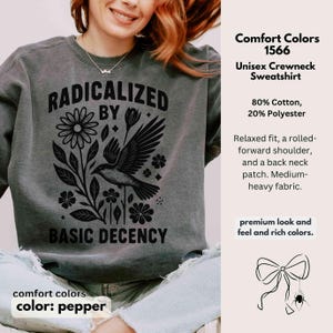 May include: Pepper-colored unisex crewneck sweatshirt with the text "RADICALIZED BY BASIC DECENCY" in black. The design includes a bird, flowers, and leaves. The sweatshirt is made of 80% cotton and 20% polyester.