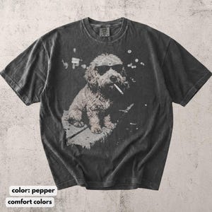 May include: Pepper-colored t-shirt featuring a graphic of a dog wearing sunglasses and smoking a cigarette. The dog is sitting down. The shirt has a vintage, distressed look. The text "comfort colors" is at the bottom of the image.