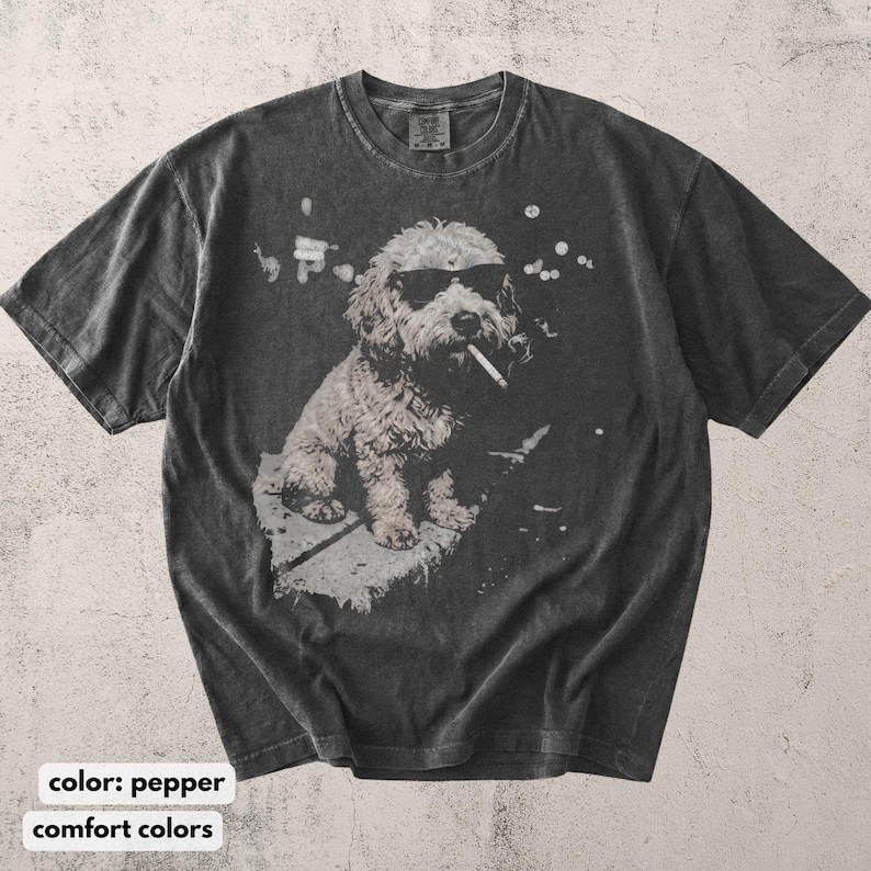 May include: Pepper-colored t-shirt featuring a graphic of a dog wearing sunglasses and smoking a cigarette. The dog is sitting down. The shirt has a vintage, distressed look. The text "comfort colors" is at the bottom of the image.