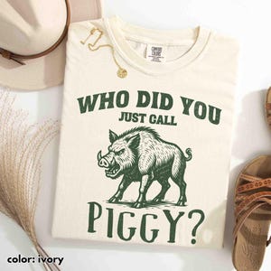 Op de afbeelding: Ivoor kleurig t-shirt met de tekst "WHO DID YOU JUST CALL PIGGY?" en een groene illustratie van een wild zwijn. Het shirt maakt deel uit van een kledingcollectie.