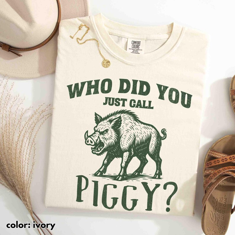 Op de afbeelding: Ivoor kleurig t-shirt met de tekst "WHO DID YOU JUST CALL PIGGY?" en een groene illustratie van een wild zwijn. Het shirt maakt deel uit van een kledingcollectie.