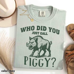 Op de afbeelding: Lichtgroen t-shirt met de tekst "WHO DID YOU JUST CALL PIGGY?" en een afbeelding van een wild zwijn. Het shirt ligt naast een hoed, sandalen en een gouden ketting.