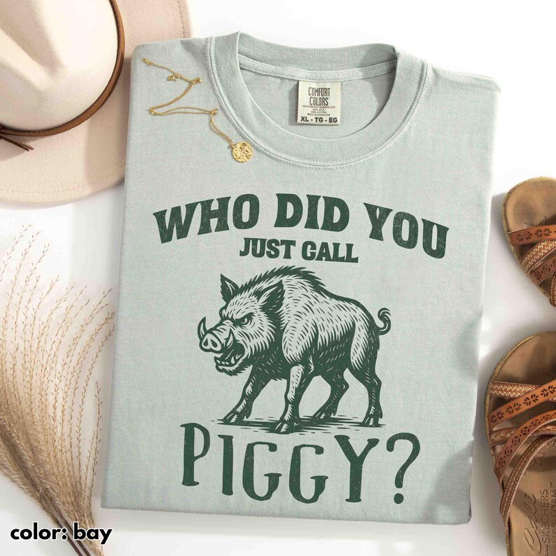 Op de afbeelding: Lichtgroen t-shirt met de tekst "WHO DID YOU JUST CALL PIGGY?" en een afbeelding van een wild zwijn. Het shirt ligt naast een hoed, sandalen en een gouden ketting.