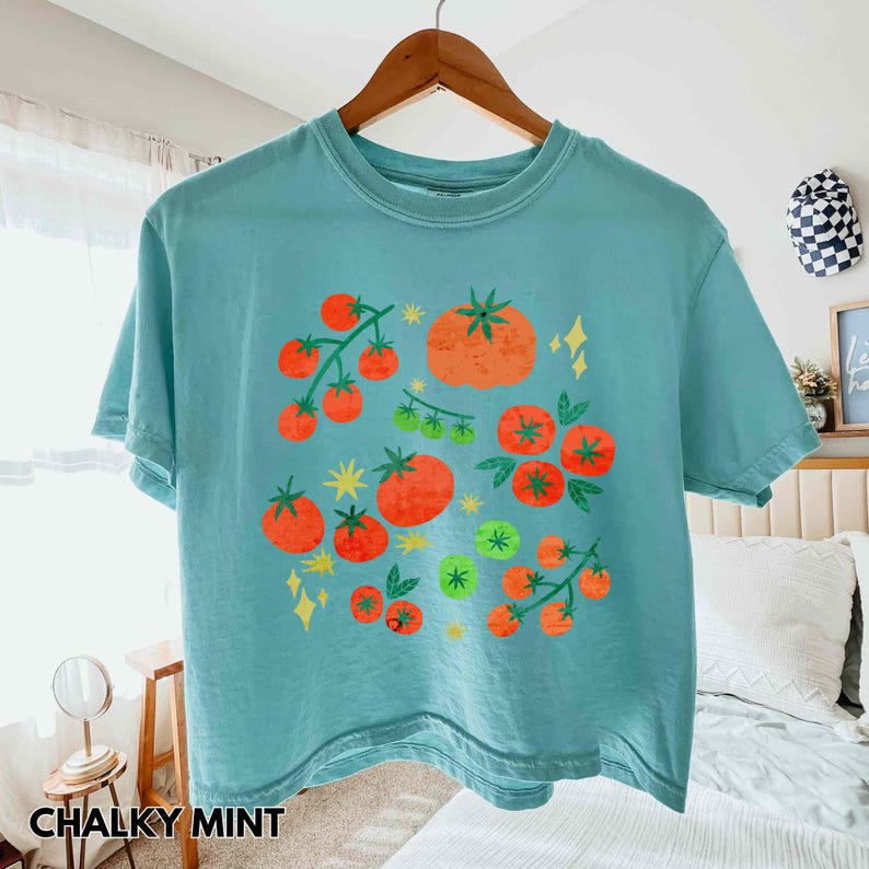 Puede incluir: Una camiseta turquesa con un dise&ntilde;o de tomate. El dise&ntilde;o presenta tomates naranjas y rojos, hojas verdes y destellos amarillos. El texto "CHALKY MINT" est&aacute; en la parte inferior de la camiseta.