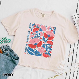 May include: Ivory-colored t-shirt featuring a floral design with red and pink flowers, blue leaves, and the text "Freedom Democracy Truth." The shirt is paired with blue jeans. The word "IVORY" is printed at the bottom.