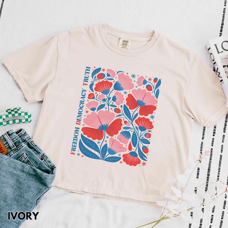 May include: Ivory-colored t-shirt featuring a floral design with red and pink flowers, blue leaves, and the text "Freedom Democracy Truth." The shirt is paired with blue jeans. The word "IVORY" is printed at the bottom.