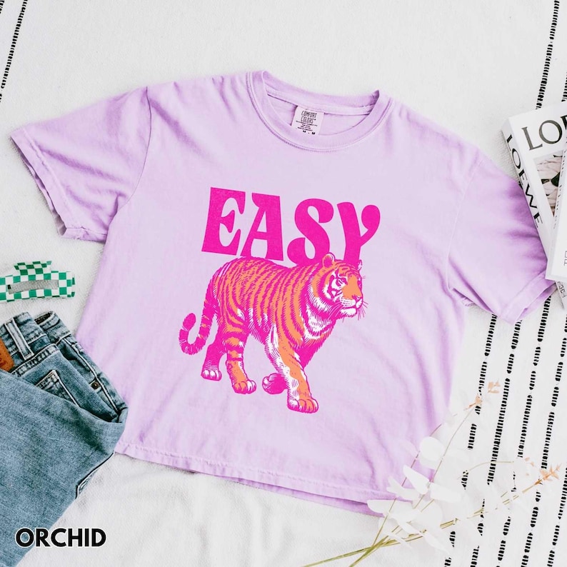 Peut inclure: T-shirt de couleur orchid&eacute;e avec le mot "EASY" en lettres roses vives. En dessous, une illustration de tigre aux tons orange et rose. Le t-shirt est associ&eacute; &agrave; un jean bleu et un petit accessoire &agrave; carreaux verts et blancs.