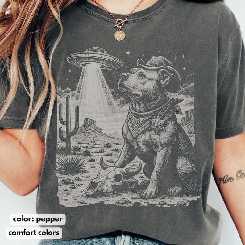 以下が含まれることがあります： コショウ色のTシャツ。カウボーイハットと保安官バッジを身に着けた犬がUFOを見上げているグラフィック。サボテン、山、牛の頭蓋骨がある砂漠の風景のデザイン。下部には「comfort colors」の文字。