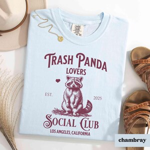 May include: Light blue t-shirt with a graphic of a raccoon and the text "Trash Panda Lovers Social Club Est. 2025 Los Angeles, California".