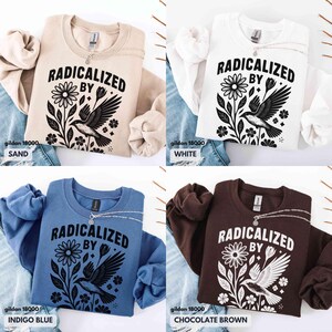 May include: Four Gildan 18000 sweatshirts in sand, white, indigo blue, and chocolate brown. Each sweatshirt features the text "RADICALIZED BY" above a floral and bird graphic in black. A silver necklace is draped on each shirt.