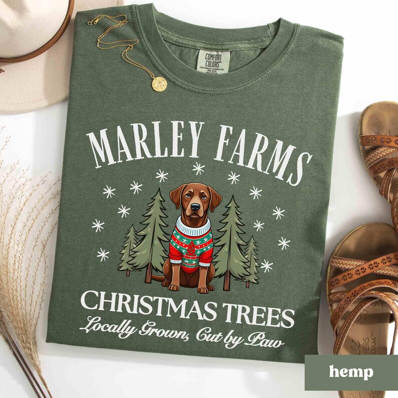 K&ouml;nnte beinhalten: Gr&uuml;nes T-Shirt mit einer wei&szlig;en Grafik eines Hundes, der einen Weihnachtspullover tr&auml;gt, und dem Text "Marley Farms Christmas Trees Locally Grown, Cut by Paw".