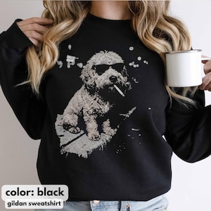 May include: Black Gildan sweatshirt featuring a graphic of a dog wearing sunglasses and smoking a cigarette. The dog is depicted in a grayscale illustration. The text "color: black" and "gildan sweatshirt" are at the bottom.