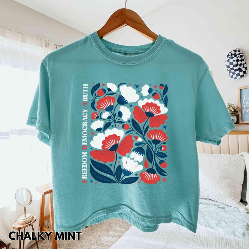 May include: A teal-colored t-shirt with a floral design featuring red and white flowers, blue leaves, and the words "FREEDOM DEMOCRACY TRUTH" printed vertically. The shirt is hanging on a wooden hanger.