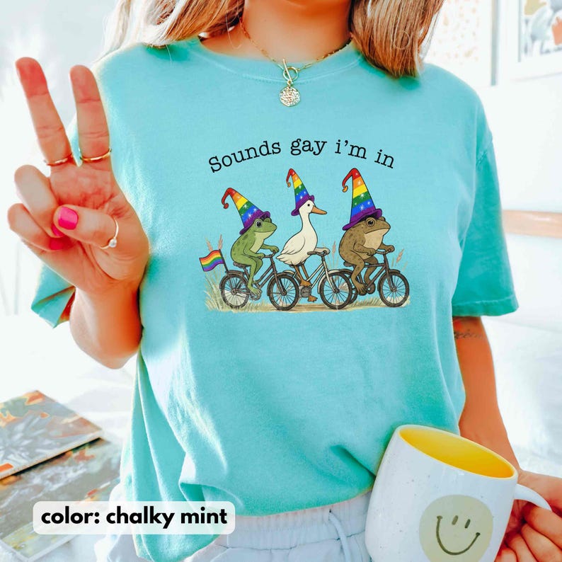 May include: Chalky mint-colored t-shirt with the phrase "Sounds gay I'm in" printed on the front. The graphic features three animals riding bicycles, each wearing a rainbow-colored wizard hat. A rainbow flag is attached to one bicycle.