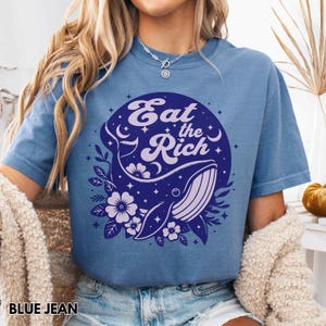 May include: A blue jean t-shirt featuring a whale and floral graphic. The graphic reads "Eat the Rich" in a retro font.