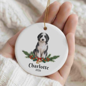 May include: A round white ceramic ornament featuring a black and white puppy illustration centered above a festive arrangement of green pine branches, red berries, and gold bells. The text Charlotte and 2026 is printed in black below the puppy.