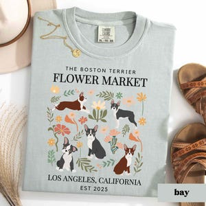 May include: Light blue t-shirt with a floral design featuring Boston Terriers. The text "The Boston Terrier Flower Market" is printed in black at the top of the shirt. The text "Los Angeles, California Est 2025" is printed in black at the bottom of the shirt.