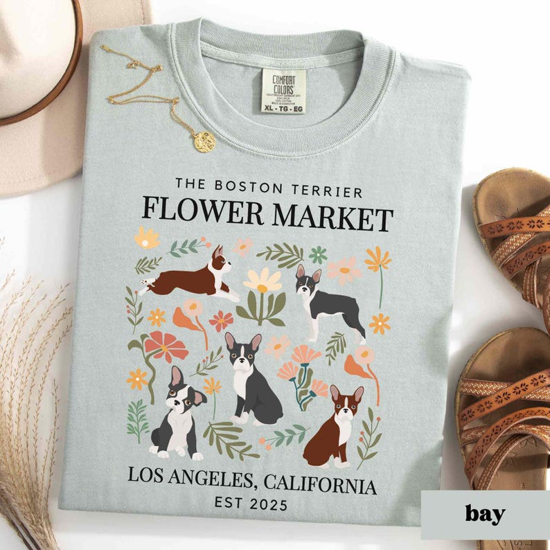 May include: Light blue t-shirt with a floral design featuring Boston Terriers. The text "The Boston Terrier Flower Market" is printed in black at the top of the shirt. The text "Los Angeles, California Est 2025" is printed in black at the bottom of the shirt.