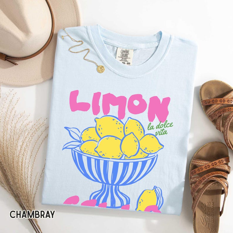 May include: Light blue t-shirt with the word "LIMON" in pink, and a bowl of yellow lemons. The text "la dolce vita" is below the bowl. A gold necklace, a hat, and sandals are also in the image.