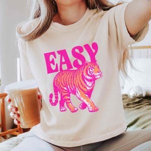 Peut inclure: T-shirt cr&egrave;me avec le mot "EASY" en lettres roses vives au-dessus d'une illustration de tigre rose et orange. Le tigre marche, la queue enroul&eacute;e. Le t-shirt est de style d&eacute;contract&eacute;.