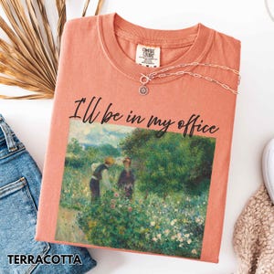 May include: A terracotta-coloured t-shirt with the text "I'll be in my office". The shirt displays a painting of two figures in a garden. A silver necklace with a sun pendant is draped on the shirt.