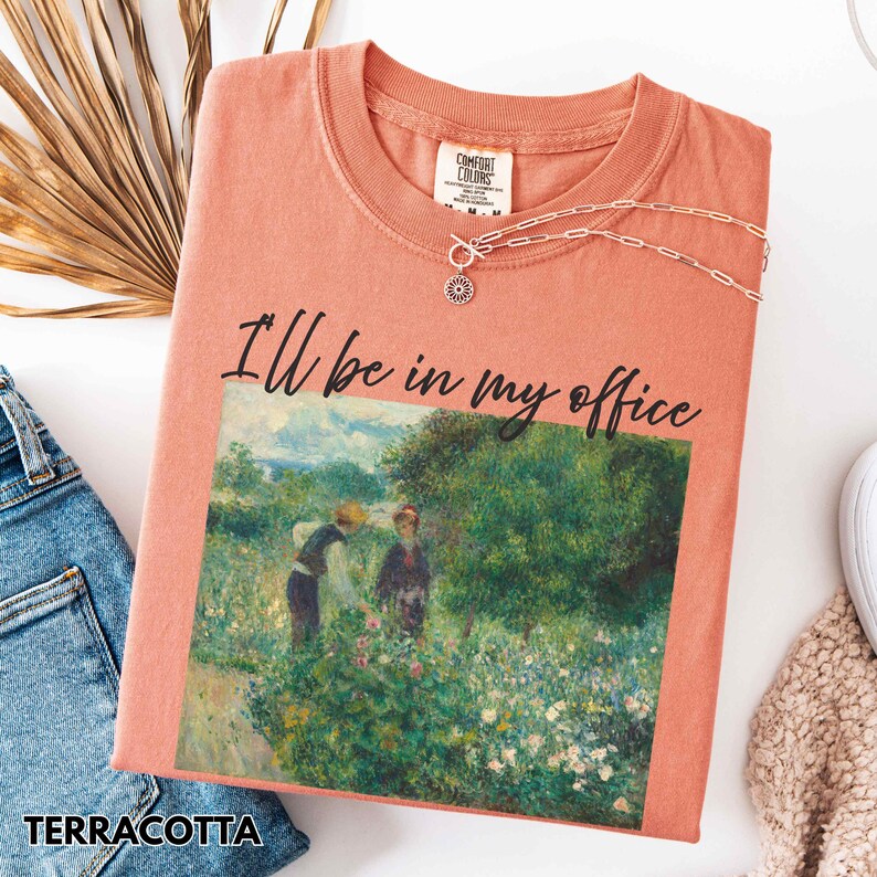 May include: A terracotta-coloured t-shirt with the text "I'll be in my office". The shirt displays a painting of two figures in a garden. A silver necklace with a sun pendant is draped on the shirt.