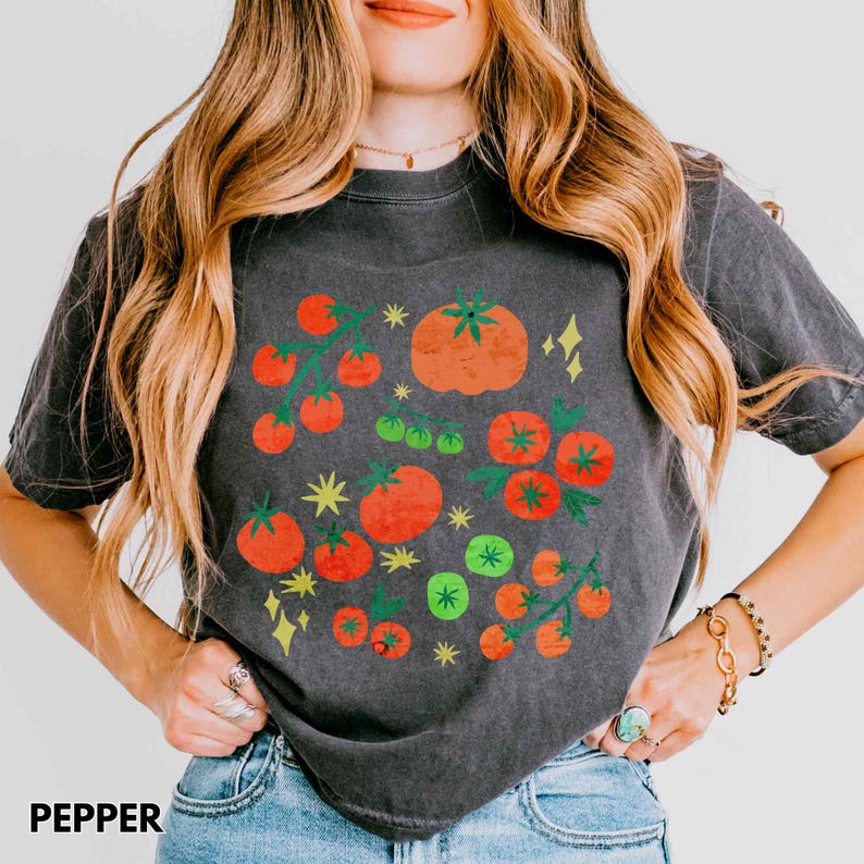 Puede incluir: Camiseta gris oscuro con un gr&aacute;fico de tomate colorido. El dise&ntilde;o presenta tomates rojos y verdes con acentos de estrellas amarillas. La palabra "PEPPER" est&aacute; impresa en la parte inferior de la camiseta.