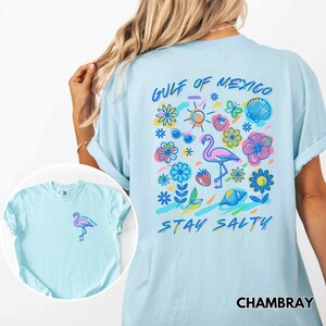 May include: A light blue t-shirt with a graphic print of a flamingo, flowers, a seashell, sunglasses, and the text "Gulf of Mexico Stay Salty". The shirt is made of a soft, comfortable fabric and has a Chambray label.