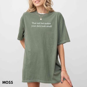 Może przedstawiać: Zielona koszulka w kolorze mchu z białym napisem: "That red hat makes your dick look small". Koszulka ma luźny kr&oacute;j i kr&oacute;tkie rękawy. Słowo "MOSS" jest wydrukowane w lewym dolnym rogu.