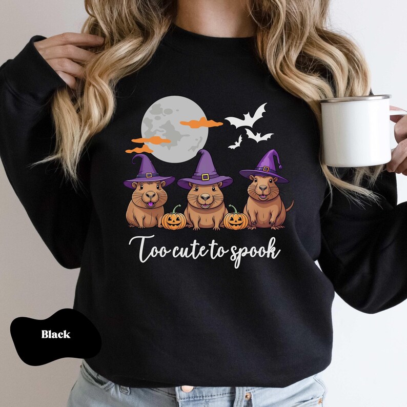 Op de afbeelding: Zwart sweatshirt met een afbeelding van drie cavia's die toverhoeden dragen en pompoenen vasthouden. De tekst "Too cute to spook" staat onder de cavia's.