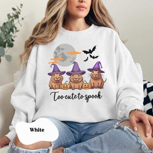 Op de afbeelding: Wit sweatshirt met een afbeelding van drie cavia's die toverhoeden dragen en pompoenen vasthouden. De tekst "Too-cute to spook" staat onder de cavia's.