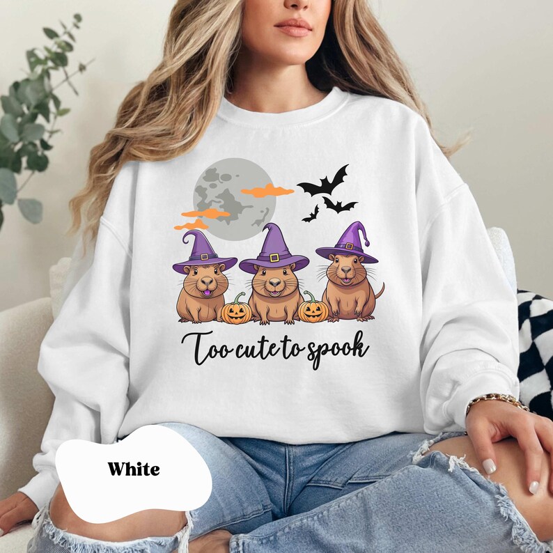 Op de afbeelding: Wit sweatshirt met een afbeelding van drie cavia's die toverhoeden dragen en pompoenen vasthouden. De tekst "Too-cute to spook" staat onder de cavia's.