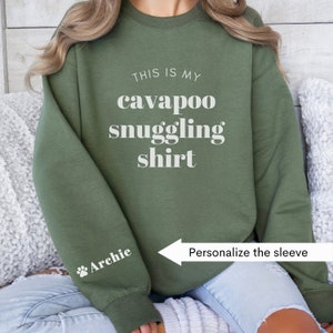 May include: A green sweatshirt with the text "This is my cavapoo snuggling shirt" printed on the front. The sleeve has a paw print and the text "Archie" printed on it.