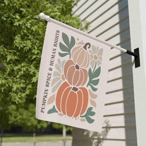 May include: A cream-colored outdoor flag featuring a stacked pumpkin design in shades of orange, brown, and beige, with green leaves and white flowers. The text "PUMPKIN SPICE & HUMAN RIGHTS" is printed vertically on the left side.