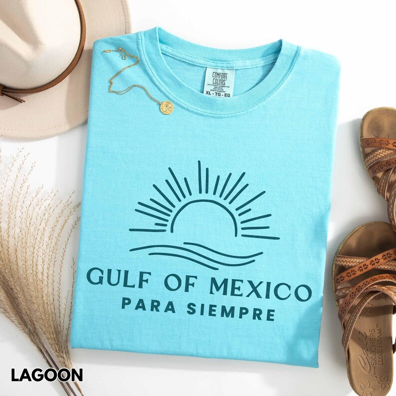 May include: A light blue t-shirt with a graphic of a sun setting over waves and the text "GULF OF MEXICO PARA SIEMPRE".