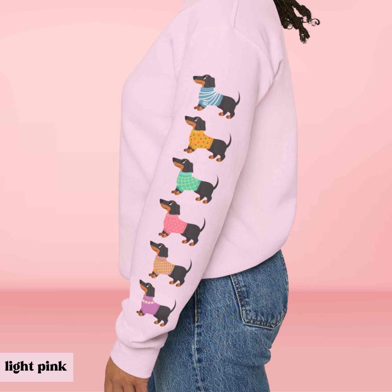 May include: A light pink sweatshirt with a row of black dachshunds wearing colorful sweaters printed on the sleeve.