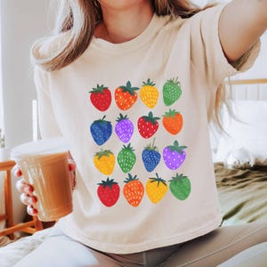May include: Cream-colored t-shirt featuring a colorful strawberry pattern. The strawberries are in various colors, including red, orange, yellow, green, blue, and purple. The shirt has a crew neck and short sleeves.