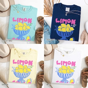 May include: Four t-shirts in different colors: chalky mint, true navy, banana, and white. Each shirt features the word "LIMON" in pink, a bowl of lemons, and the phrase "la dolce vita".