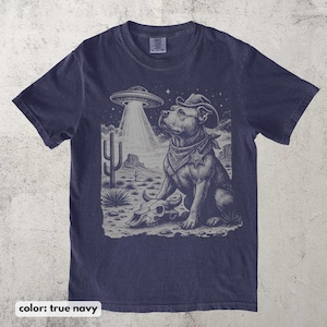 以下が含まれることがあります： ネイビーブルーのTシャツ。カウボーイハットとバンダナを身に着けた犬がUFOを見上げているグラフィック。デザインには、頭蓋骨とサボテンのある砂漠の風景が含まれています。「color: true navy」の文字が下にあります。