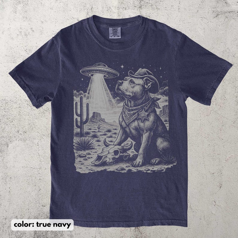 以下が含まれることがあります： ネイビーブルーのTシャツ。カウボーイハットとバンダナを身に着けた犬がUFOを見上げているグラフィック。デザインには、頭蓋骨とサボテンのある砂漠の風景が含まれています。「color: true navy」の文字が下にあります。