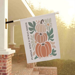 May include: A white outdoor flag with the text "PUMPKIN SPICE & HUMAN RIGHTS" in a vertical orientation. The flag features a stack of three pumpkins in shades of orange and tan, with green leaves and white flowers.
